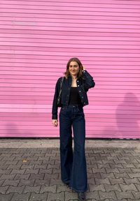 Temno modra jeans jakna in široke kavbojke z visokim pasom, združene s črnim topom. Dodatki vključujejo večplastne ogrlice in majhno torbico. Rožnata ozadje.