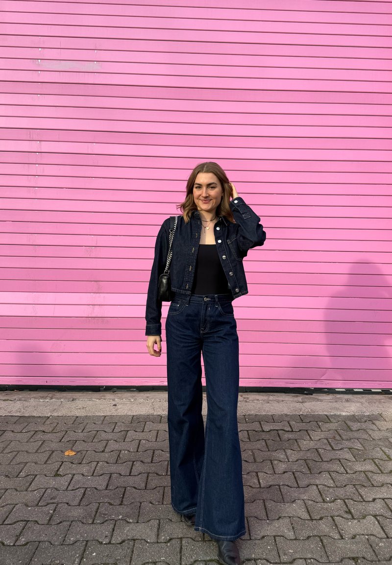 Donker denimjack en wijde, hooggetailleerde jeans gecombineerd met een zwart topje. Accessoires omvatten gelaagde kettingen en een kleine tas. Roze achtergrond.