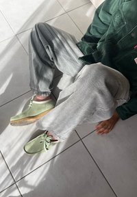 Hellgrüne Schuhe mit runden Zehen und Schnürsenkeln, graue schmal geschnittene Hose und eine dunkelgrüne Cordjacke auf gefliesten Boden mit sanften Schatten.