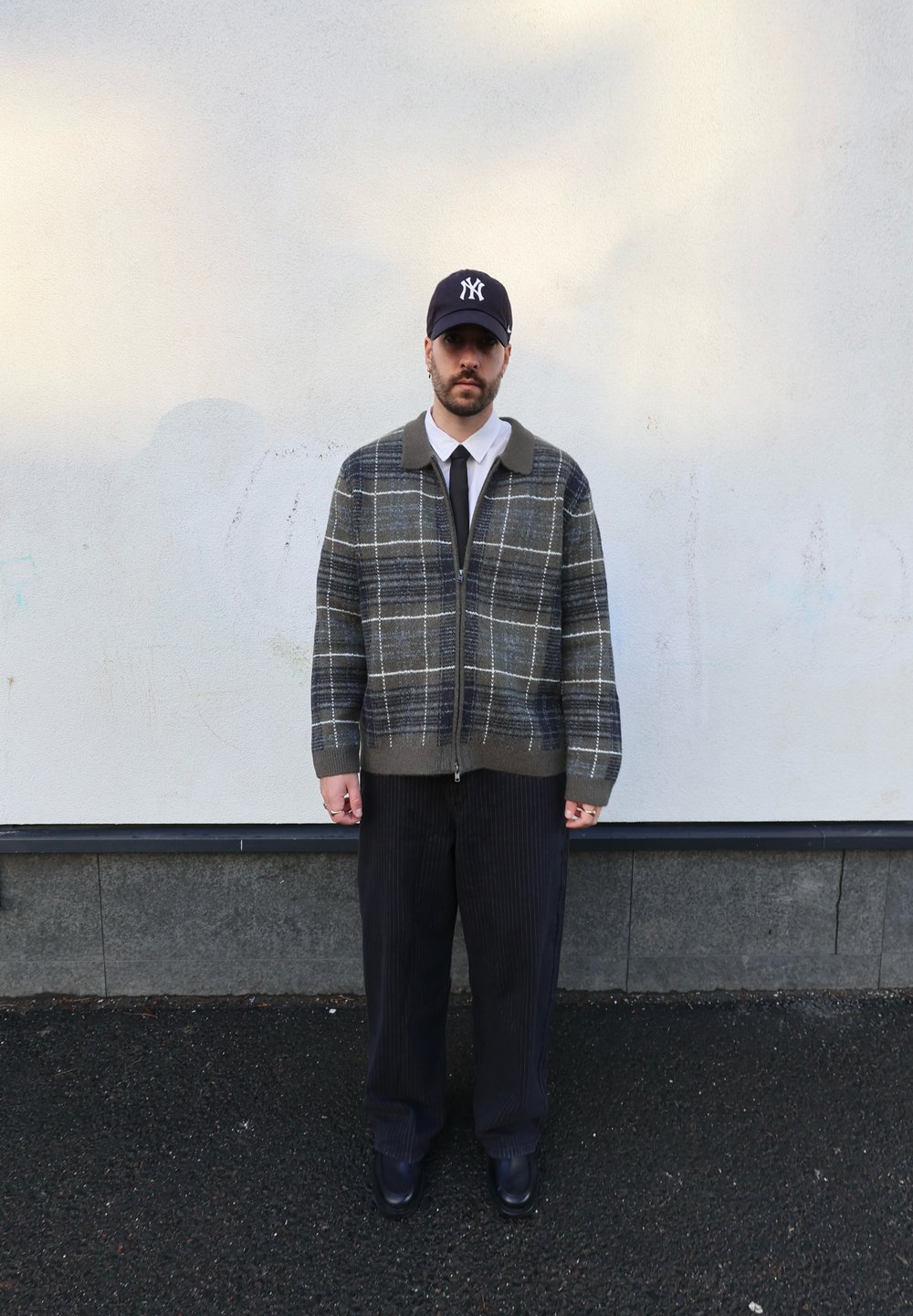 Veste zippée en plaid gris avec un col sombre, associée à un pantalon noir à fines rayures et des chaussures noires, portée avec une casquette foncée.