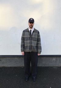 Veste zippée en plaid gris avec un col sombre, associée à un pantalon noir à fines rayures et des chaussures noires, portée avec une casquette foncée.