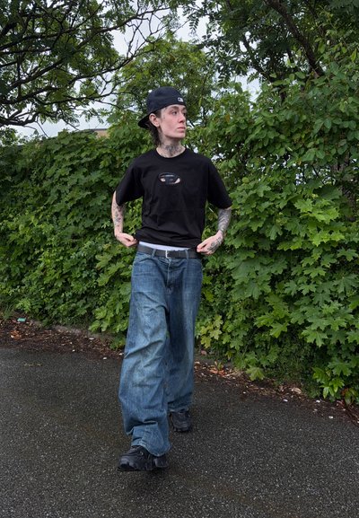 Persona con tatuajes que lleva una gorra negra al revés, camiseta negra y vaqueros anchos azules caminando por un camino de asfalto mojado junto a arbustos verdes.