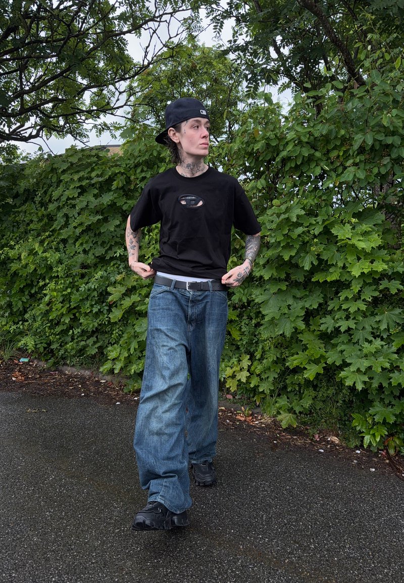 Personne avec des tatouages portant une casquette noire à l'envers, un t-shirt noir et un jean bleu ample marchant sur un chemin en asphalte mouillé à côté d'arbustes verts.