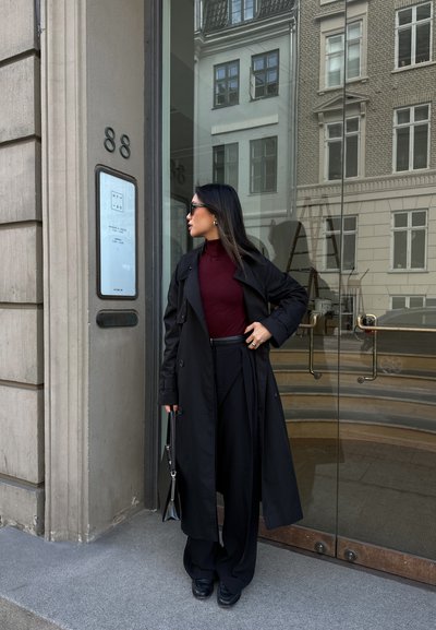 Abrigo negro sobre un suéter de cuello alto burdeos, combinado con pantalones de pierna ancha negros. Los accesorios incluyen gafas de sol oscuras y un bolso negro pequeño.