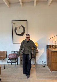 Femme portant des lunettes, une veste olive ceinturée, un pantalon noir et des bottes, debout dans une pièce moderne avec des œuvres d'art abstrait, des meubles en bois et des bougies allumées.