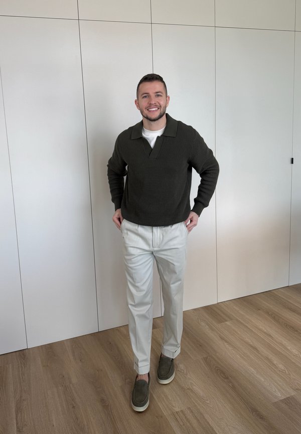 Uśmiechnięty mężczyzna w ciemnozielonym swetrze, białych spodniach i zielonych butach wsuwanych stojący na drewnianej podłodze przed białą ścianą z paneli.