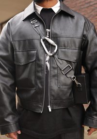 Veste en cuir noire avec une fermeture éclair à l'avant, deux poches poitrine et des accessoires métalliques. Associée à un t-shirt graphique noir et un petit sac.