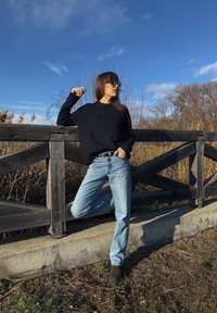 Γυναίκα με μαύρο πουλόβερ και γυαλιά ηλίου στηρίζεται σε ξύλινο φράχτη, με το ένα πόδι σηκωμένο, σε εξωτερικό χώρο κάτω από καθαρό γαλάζιο ουρανό με φόντο ξερά φύλλα.