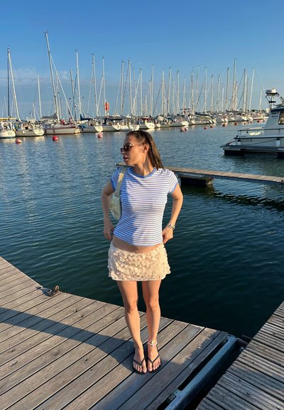 Camisa de rayas de manga corta, falda corta de volantes beige, chanclas negras y un bolso de color claro. Muelle de madera y marina al fondo.