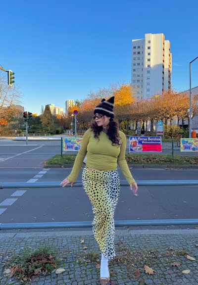 Top de manga larga verde oliva combinado con una falda amarilla con manchas negras, posando en un entorno urbano bajo un cielo azul.