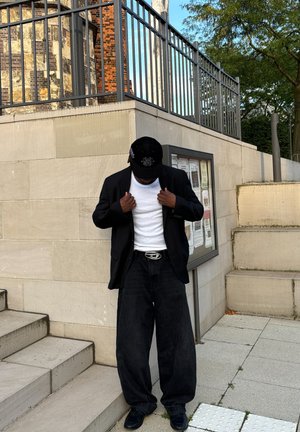 Blazer negro sobre una camiseta blanca, combinado con unos vaqueros negros de corte holgado y zapatos negros. Una gorra negra completa el conjunto.