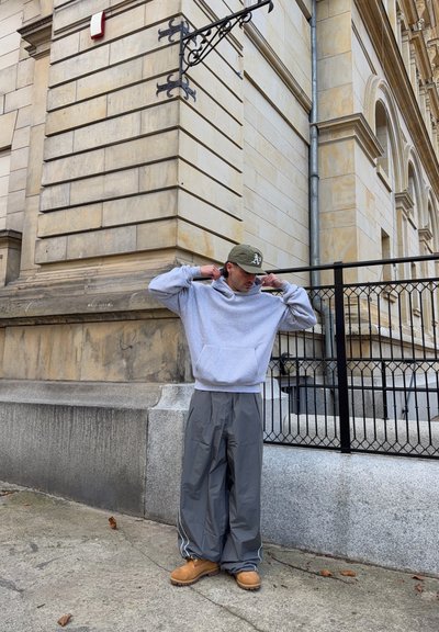 Sudadera gris con bolsillo frontal, pantalones grises de pierna ancha con rayas laterales blancas y botas marrones. Al fondo, un edificio de piedra y una cerca.