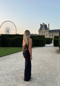 Musta, vartalonmyötäinen pitkä hame yhdistettynä ruskeaan, pilkulliseen puseroon. Taustalla vihreitä pensasaitoja ja historiallinen rakennus.