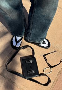 Zwarte sandalen met gekruiste banden, witte sokken, donkere denim broek. In de buurt: een zwarte leren tas met gespen, zonnebril en een parelarmband.
