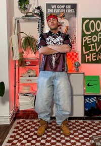 Homme en jean ample, t-shirt graphique, bandana rouge et bottes, debout les bras croisés dans une pièce avec des baskets, des plantes et des affiches.