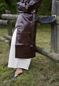 Marron brillant manteau en cuir synthétique avec une ceinture à nouer, associé à une jupe midi texturée blanche et des chaussures noires plates, appuyé contre une clôture en bois.