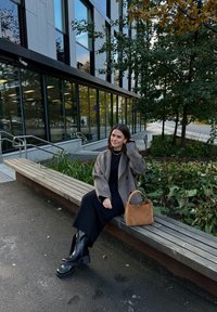 Hnedý oversized kabát, čierne rebrované šaty, masívne čierne čižmy. Hnedá kabelka na lavičke, drevené sedenie, zelené rastliny, moderná budova v pozadí.