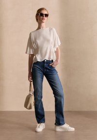 Top bianco con texture e maniche corte, abbinato a jeans in denim blu. Il modello indossa sneakers bianche e tiene in mano una piccola borsa beige. Sfondo marrone.
