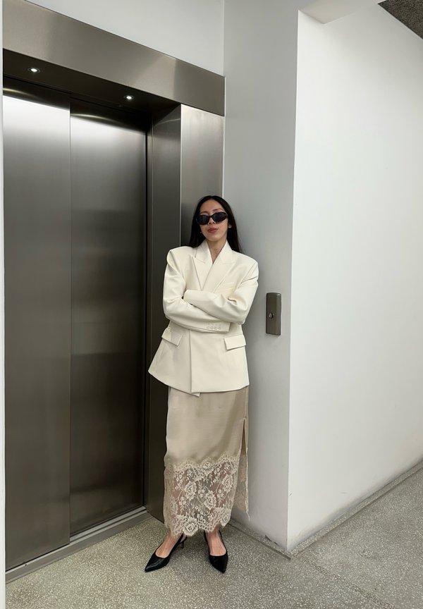 Kvinne iført hvit blazer og kremfarget blondeskjørt står med armene i kors nær lukkede heisdører i en moderne, minimalistisk gang.