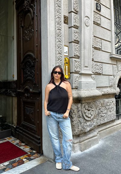 Top corto negro con cuello halter, jeans descoloridos de un azul claro y zapatos deslizantes beige. De pie junto a una entrada de piedra y madera con texturas intrincadas.