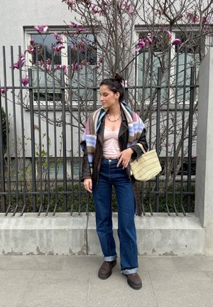 Mujer de pie en la acera cerca de una valla metálica negra y un árbol con flores rosas, lleva chaqueta de cuero, vaqueros, bufanda a rayas y sostiene un bolso de paja.