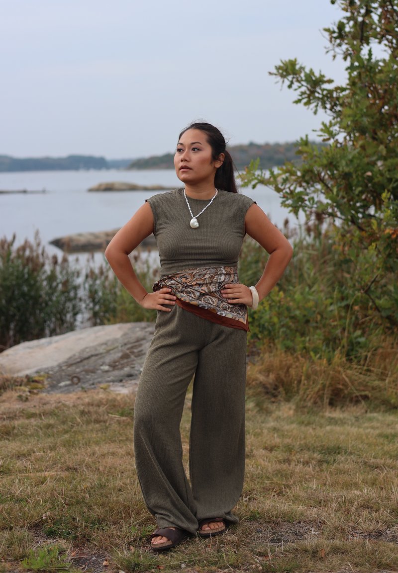 Woman standing outdoors near water, wearing olive green sleeveless top and trousers, with a patterned scarf tied around her waist and a shell necklace.