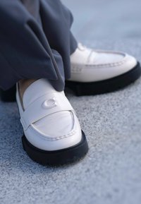 Λευκά δερμάτινα loafers με μαύρη καουτσούκ σόλα, διαθέτοντας μπροστινή λωρίδα και ανάγλυφο λογότυπο. Λείο υφή με ελαφριά γυαλάδα.