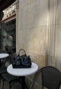 Čierna kožená kabelka s dvoma hornými úchytmi, striebornými doplnkami a detailmi z pracky, leží na okrúhlom bielom stole oproti kamennej stene.