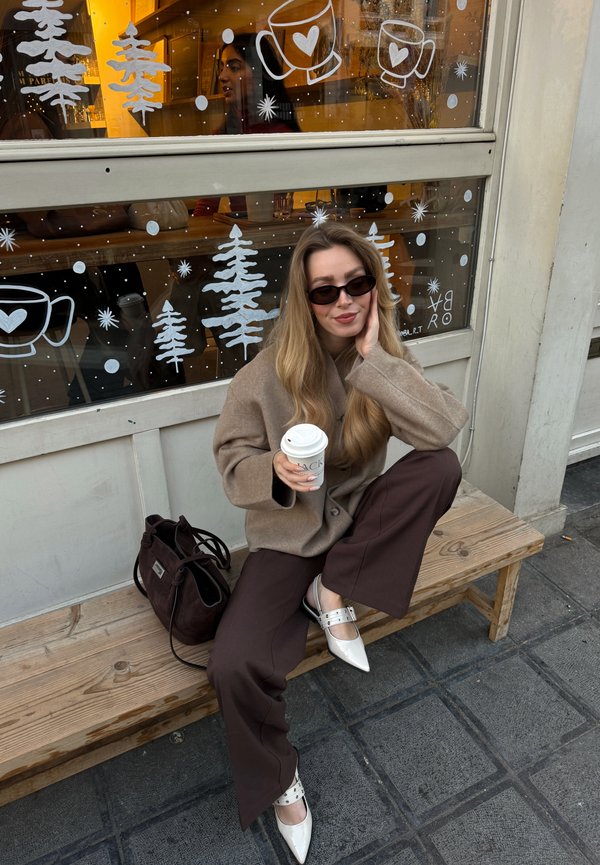 Joven mujer con gafas de sol y abrigo beige sentada en un banco de madera fuera de una cafetería, sosteniendo una taza de café, con dibujos de temática invernal en la ventana detrás de ella.