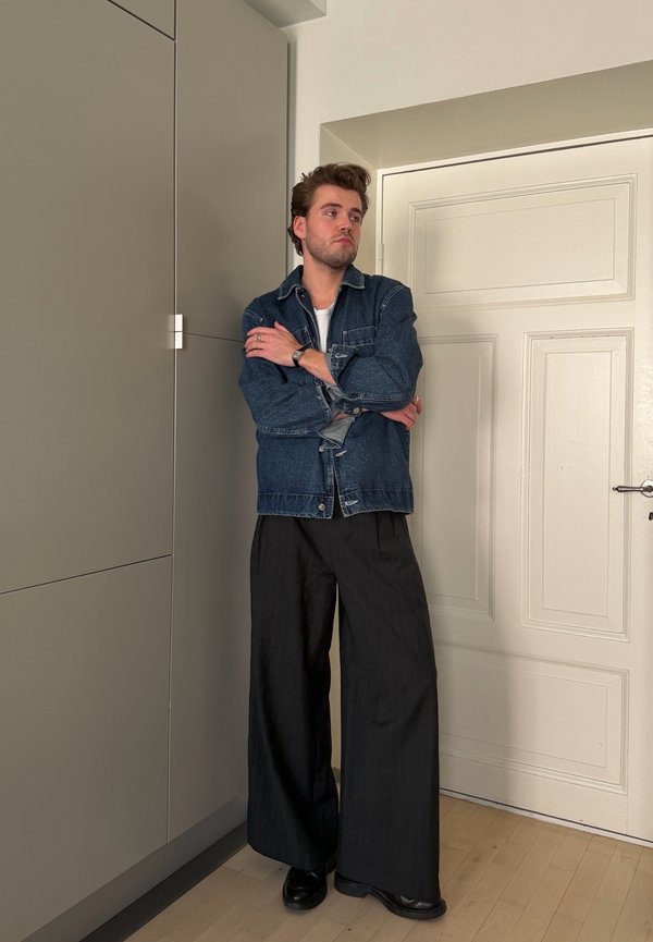 Giovane uomo con pantaloni neri larghi e giacca di jeans in piedi con le braccia incrociate vicino a una porta bianca e armadietti grigi all'interno.