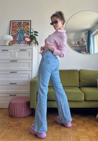 Camisa listrada em rosa claro e branco, jeans flare de cintura alta em azul claro, Crocs lilás, sofá verde, espelho redondo, arte de parede colorida.