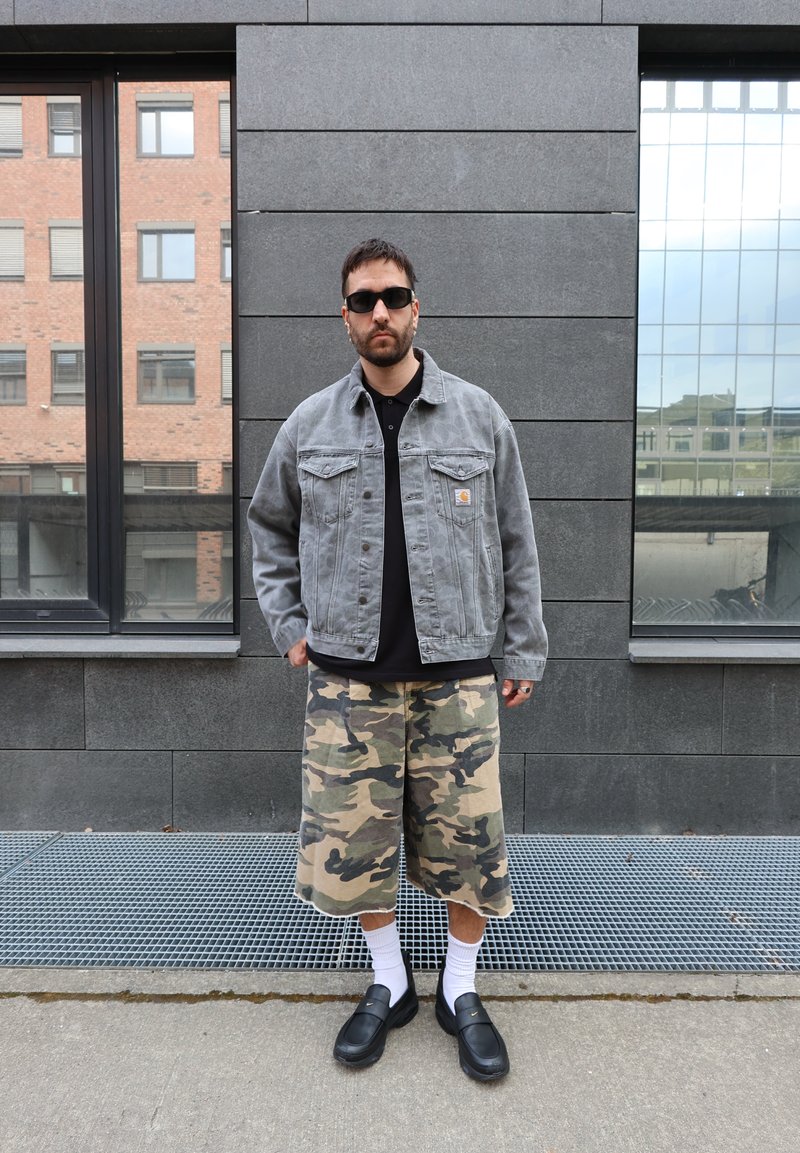 Homme portant des lunettes de soleil, une veste en jean grise, une chemise noire, un short camouflage, des chaussettes blanches et des mocassins noirs se tient dehors devant un mur gris.