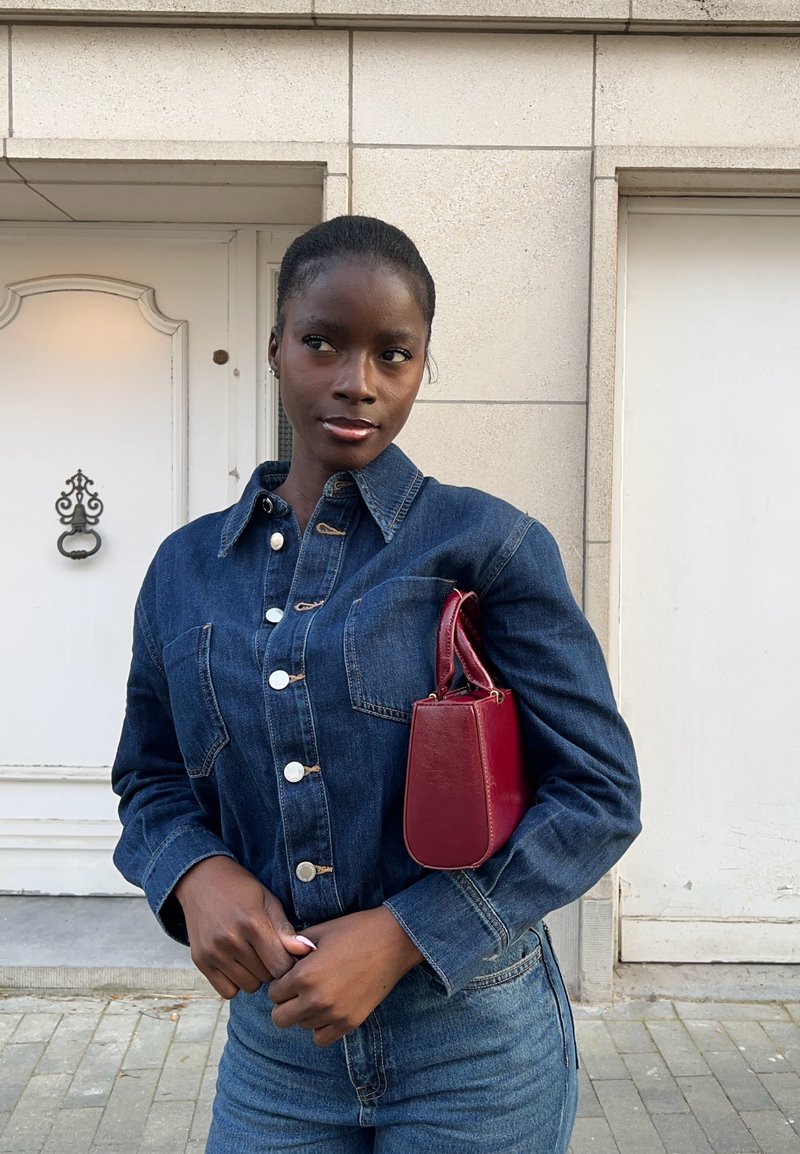 Jeune femme à la peau foncée portant une veste en jean et un jean, tenant un petit sac à main rouge, debout devant un mur en pierre avec des portes.
