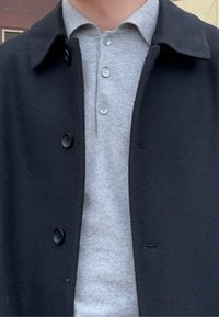 Άτομο που φοράει γκρι πόλο πουκάμισο με κουμπιά κάτω από μαύρο μάλλινο παλτό με μεγάλα κουμπιά, ορατό από τον λαιμό έως το μέσο του κορμού.