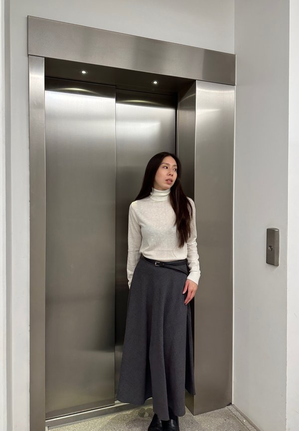 Des portes d'ascenseur en acier inoxydable, avec une personne en pull col montant clair et en jupe noire, debout à côté de l'ascenseur, sa main reposant sur sa hanche.