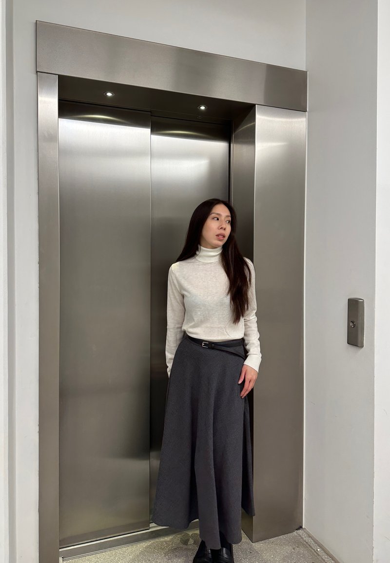 Kovinske vrata dvigala iz nerjavečega jekla, ob katerih stoji oseba v svetlem puloverju z visokim ovratnikom in temni krilu, roka položena na boku.