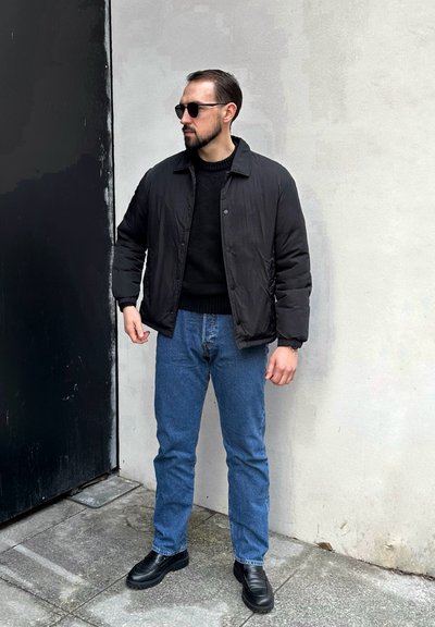 Chaqueta de plumas negra sobre un suéter negro, combinada con jeans azules y zapatos negros. Lleva gafas de sol. De pie contra un fondo neutral.