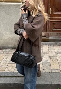 Abrigo marrón a cuadros con cuello alto, mangas largas grises y un bolso negro con relieve de cocodrilo. Jeans azules desgastados y zapatos marrones.