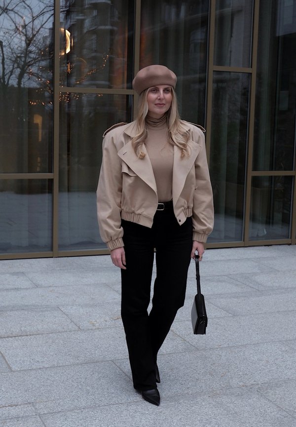 Blouson aviateur beige avec manches bouffantes et accents aux épaules, porté avec un col roulé beige et un pantalon noir évasé, tenant un petit sac noir.