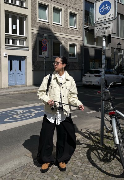 Persona joven con chaqueta beige y pantalones negros sosteniendo una cámara, de pie junto a una bicicleta en una calle empedrada con carril bici y señales.