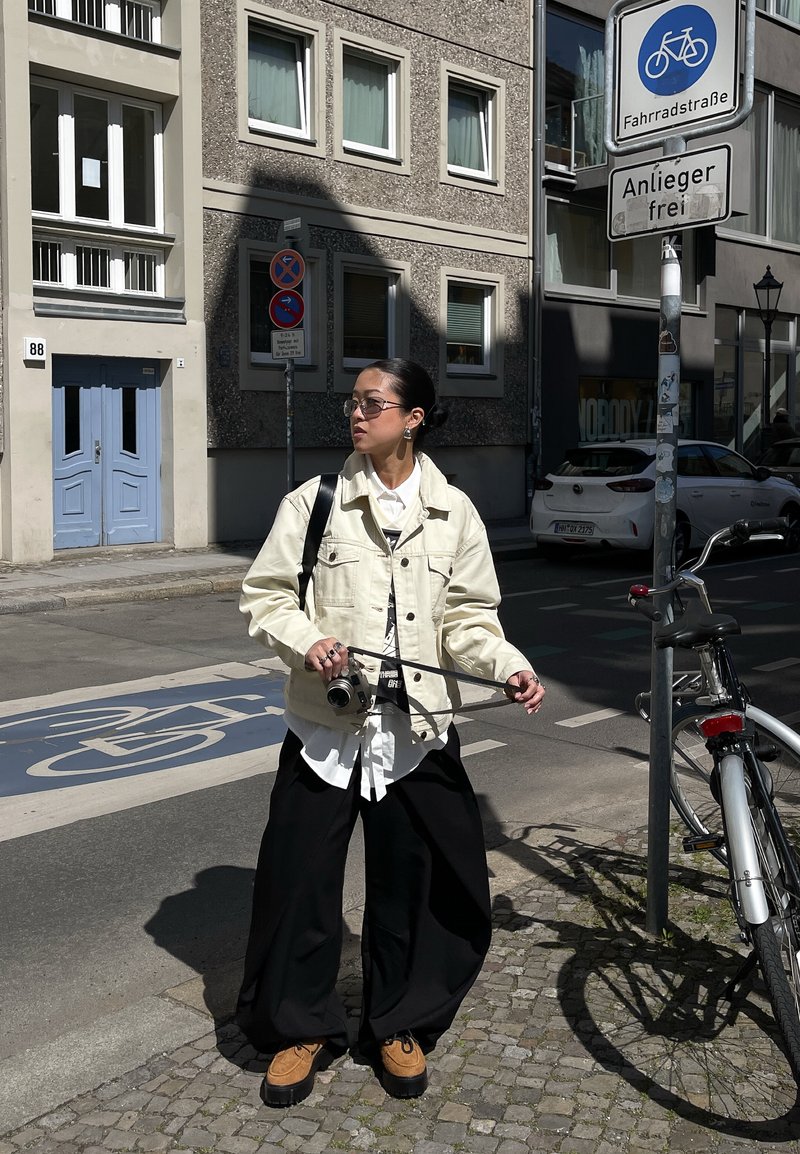 Jeune personne en veste beige et pantalon noir tenant un appareil photo, debout à côté d’un vélo dans une rue pavée avec une piste cyclable et des panneaux.