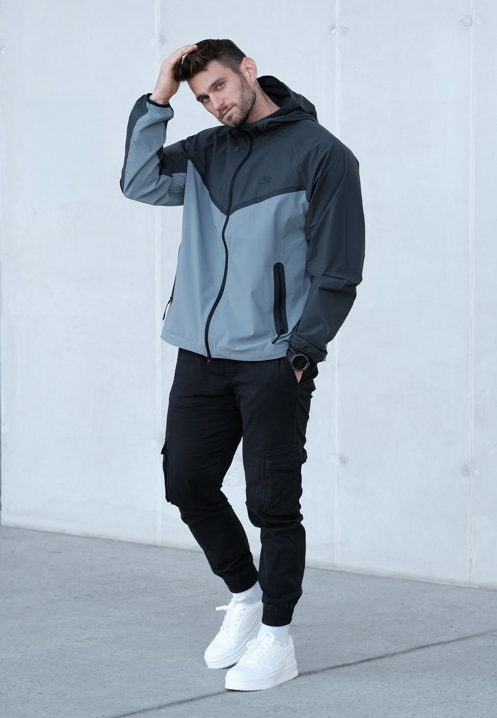 Męska lekka kurtka w ciemnoszarym i jasnoszarym kolorze, z kapturem, przednim zamkiem błyskawicznym i dwoma bocznymi kieszeniami, noszona z czarnymi spodniami typu cargo i białymi tenisówkami.
