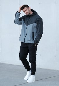 Męska lekka kurtka w ciemnoszarym i jasnoszarym kolorze, z kapturem, przednim zamkiem błyskawicznym i dwoma bocznymi kieszeniami, noszona z czarnymi spodniami typu cargo i białymi tenisówkami.