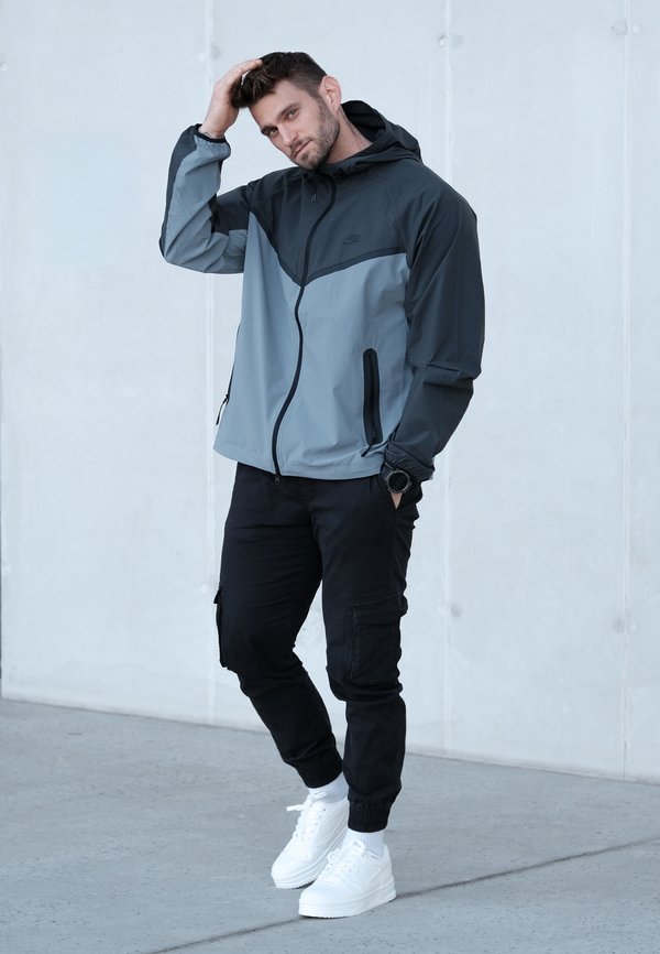 Chaqueta ligera de hombre en gris oscuro y gris claro, con capucha, cremallera frontal y dos bolsillos laterales, combinada con pantalones cargo negros y zapatillas blancas.