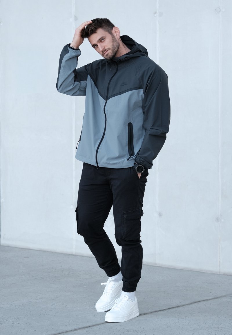 Veste légère pour hommes en gris foncé et gris clair, avec capuche, fermeture éclair frontale et deux poches latérales, portée avec un pantalon cargo noir et des baskets blanches.