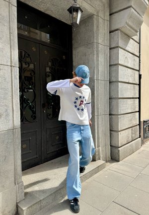 Persona con gorra azul, camisa blanca de manga larga con estrellas y una "P", vaqueros azul claro y zapatos negros bajando de un marco de piedra.