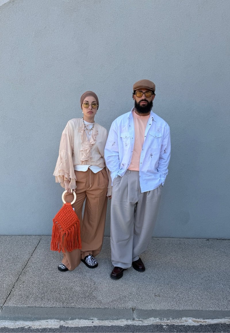 Un couple se tient contre un mur gris ; la femme porte un foulard beige, un chemisier à volants, un pantalon marron avec un sac à franges orange ; l'homme porte une casquette, une chemise blanche et un pantalon gris.