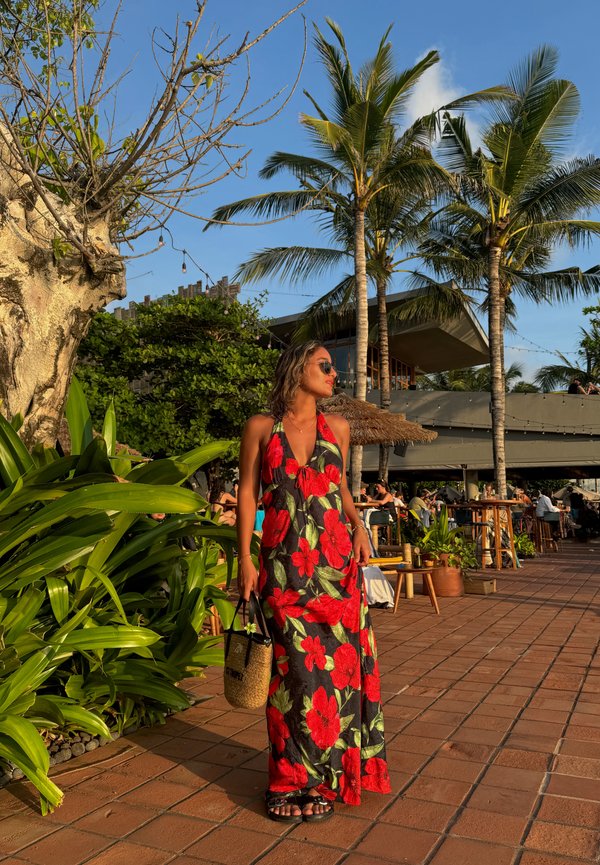 Rochie maxi cu fundal negru și imprimeu floral mare roșu. Purtați ochelari de soare, ținând o geantă țesută, stând pe o terasă exterioară cu plăci.