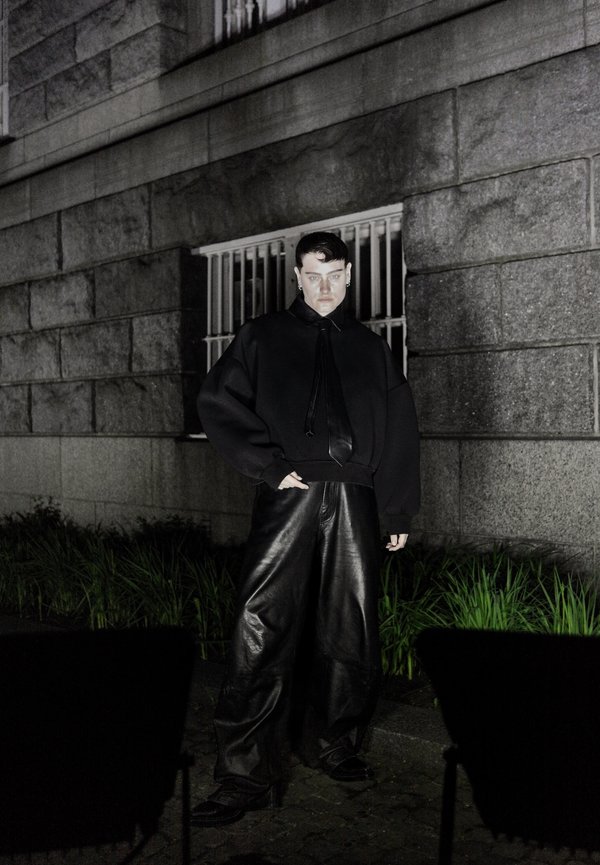 Czarny oversize hoodie z detalem w postaci wiązania, zestawiony z szerokimi czarnymi skórzanymi spodniami, na tle kamiennej ściany w słabym świetle.