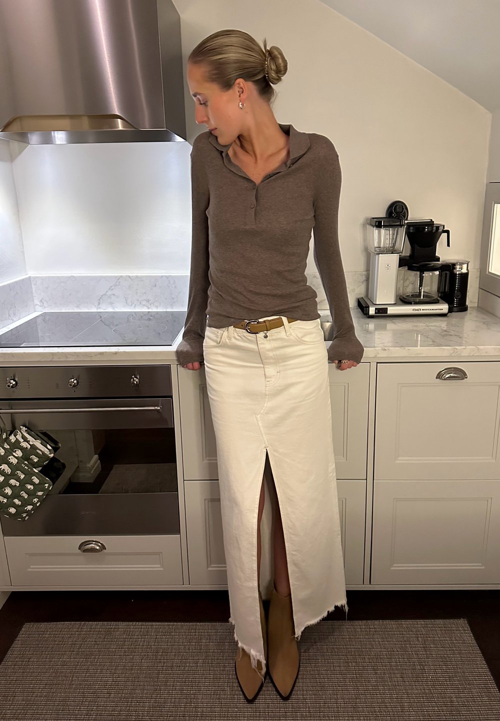 Καφέ μακρυμάνικο πόλο πουκάμισο, λευκή μέγιστη φούστα με μπροστινή σχισμή και ακατέργαστο τελ είωμα, ταμπά μπότες με μυτερή μύτη μέχρι τον αστράγαλο, σύγχρονη κουζίνα στο φόντο.